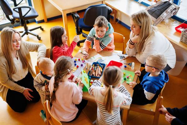 Blick auf einen Tisch mit spielenden Kindern. Es sitzt noch eine Erzieherin mit am Tisch. Eine weitere Erzieherin kniet auf dem Teppich.